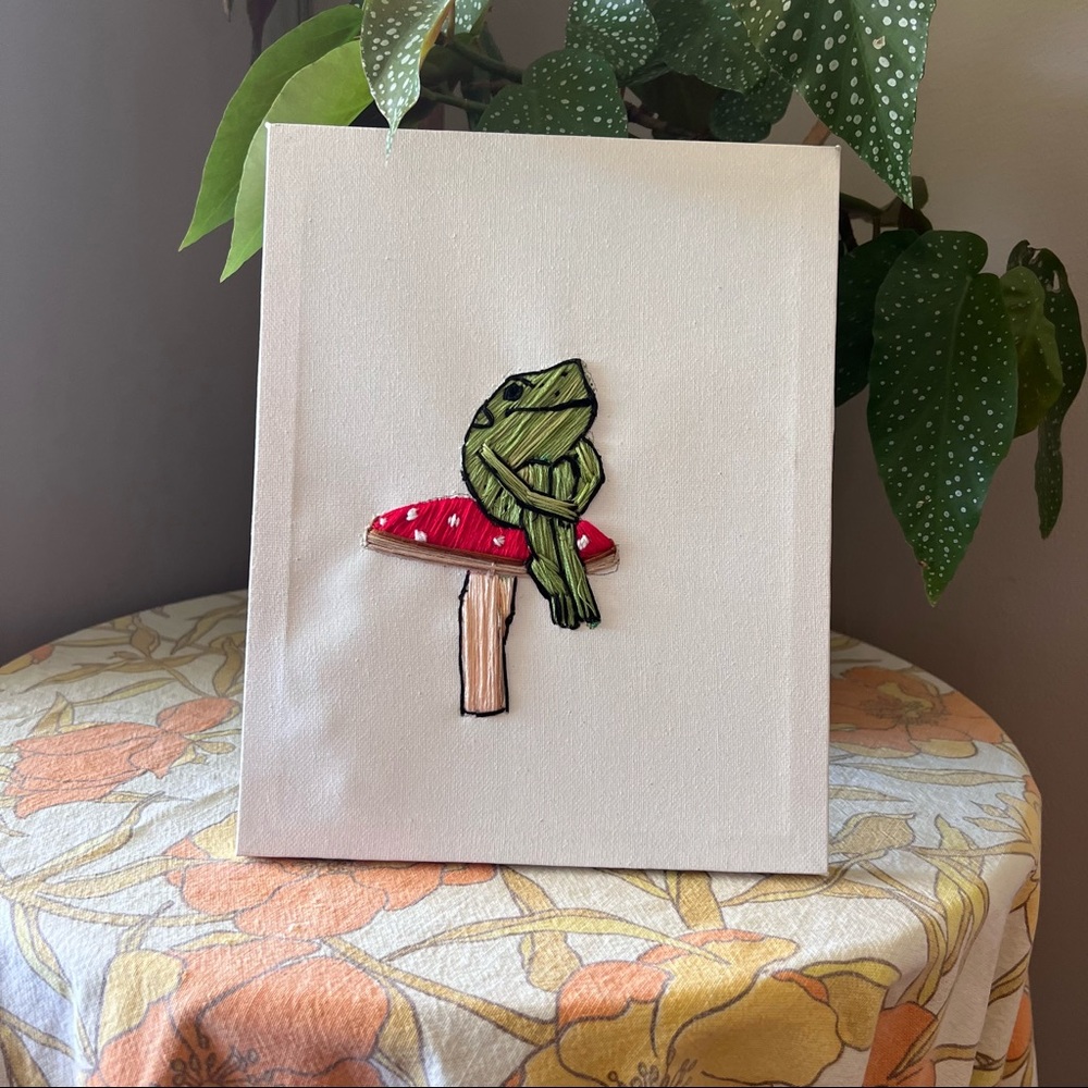 Canvas hand embroidery of a frog sitting on a mushroom